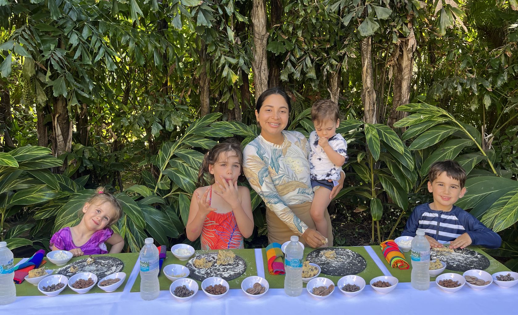 A We Care caregiver and four children at an outdoor table making cookies together, surrounded by tropical foliage in Punta Mita